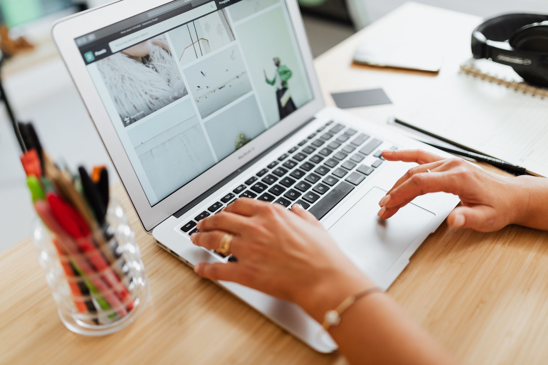 Hands using a laptop on a wooden desk with stationery nearby, browsing images online.